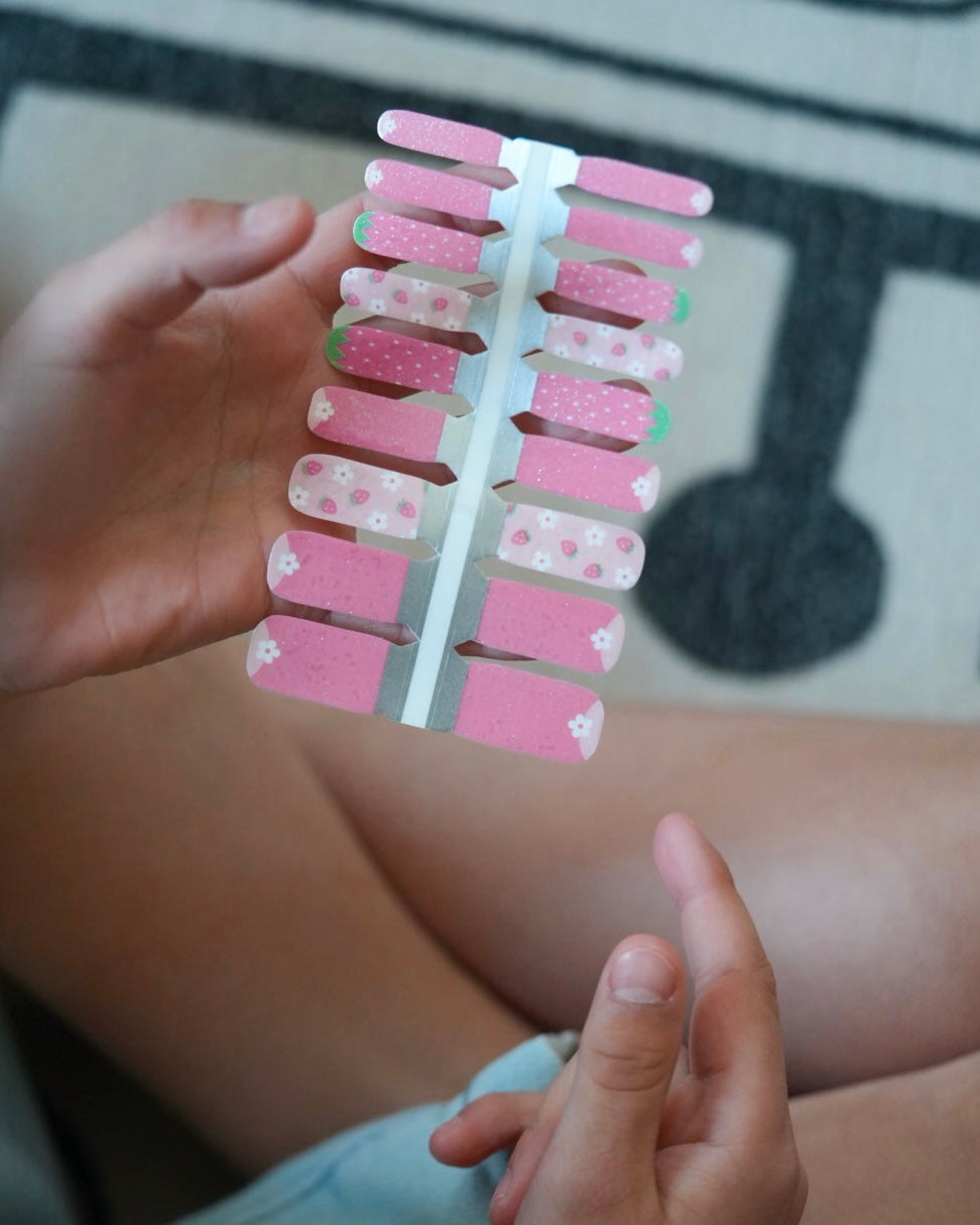 Nahaufnahme einer Kinderhand mit den Sheesh Nails Kinder-Nagelfolien „Pink Berry“. Die selbstklebenden Sticker zeigen ein zartes Rosa mit weißen Blumen und kleinen Beerenmotiven – verspielt, fröhlich und kindgerecht. Die Folien sind vollständig deckend, lassen sich leicht aufkleben und rückstandsfrei wieder entfernen – ganz ohne UV-Lampe oder zusätzliche Hilfsmittel. Hautfreundlich, ungiftig und perfekt für kleine Nail-Artists, die Farbe und Kreativität lieben.