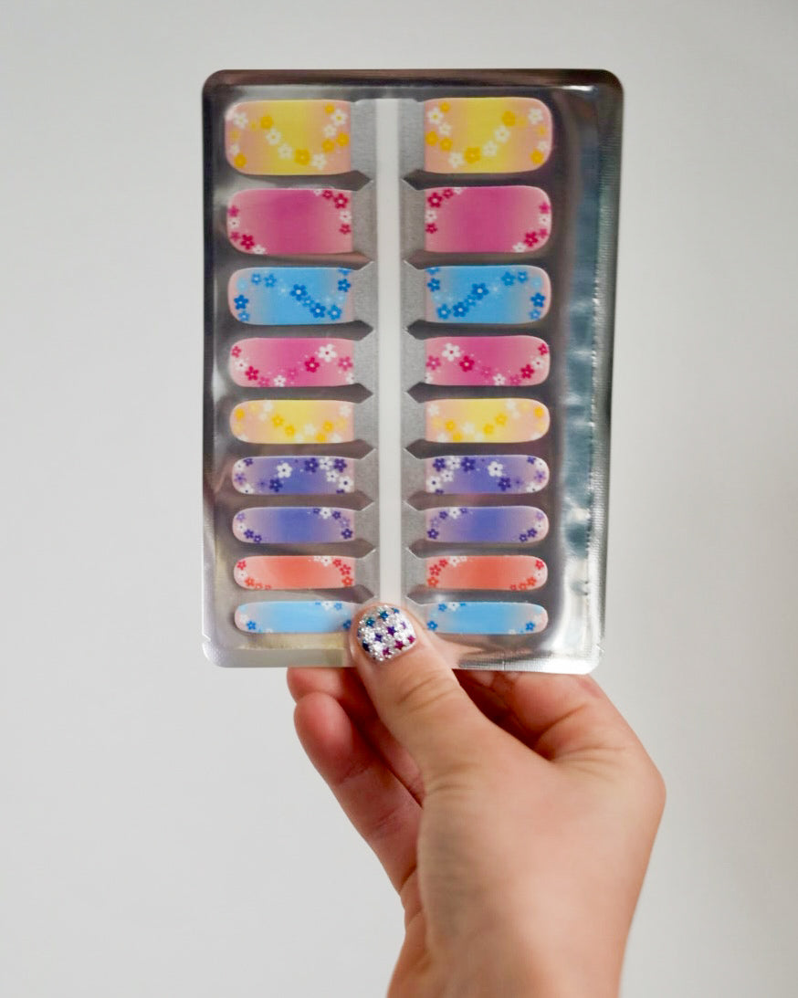 Kinderhand hält SHEESH Nails Nagelfolien Rainbow Blossom mit bunten Blumen in Pastellfarben – fröhliches Nageldesign für Kinder mit Regenbogen- und Blütenmuster.