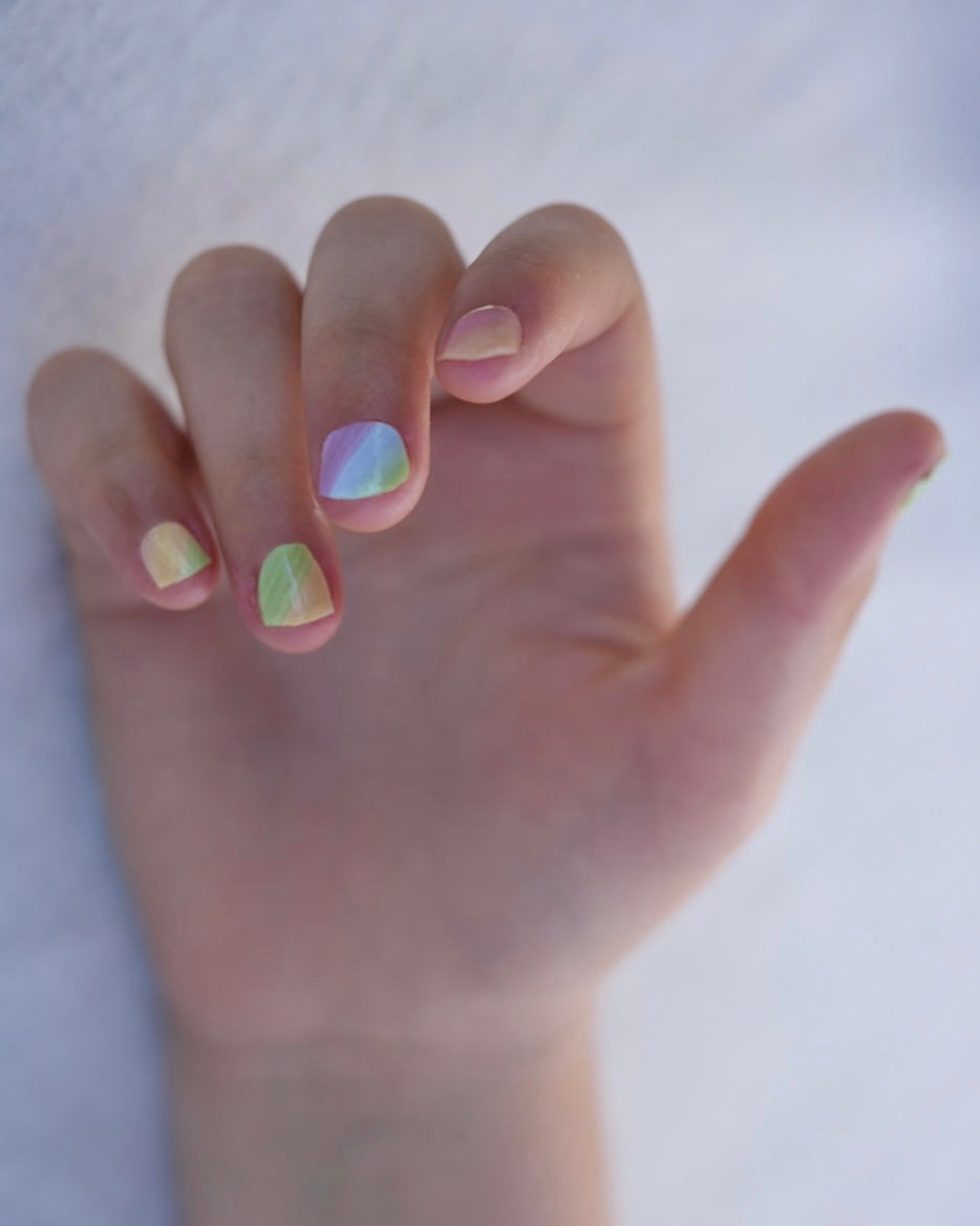 Nahaufnahme einer Kinderhand mit Sheesh Nails Kinder-Nagelfolien „Rainbow“. Die selbstklebenden Sticker zeigen ein fröhliches Regenbogendesign in sanften Pastelltönen. Das deckende Finish sorgt für einen gleichmäßigen, farbintensiven Look. Die Folien sind leicht anzubringen, hautfreundlich, ungiftig und lassen sich rückstandsfrei wieder entfernen – ideal für kreative Kinder-Maniküren mit Farbe und Spaß.