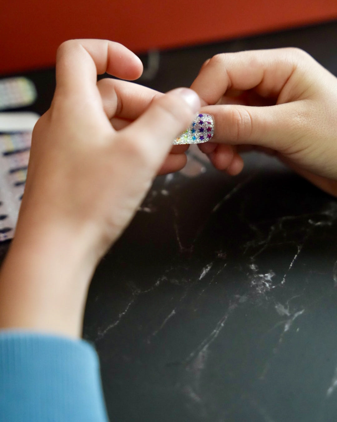 Kinderhand hält bunte Glitzer-Nagelfolien mit Regenbogen-Sternchen von SHEESH Nails – fröhliches Naildesign für Kinder mit funkelndem Regenbogenmuster.
