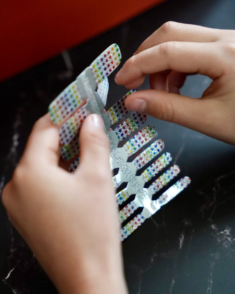 Kinderhand hält bunte Glitzer-Nagelfolien mit Regenbogen-Sternchen von SHEESH Nails – fröhliches Naildesign für Kinder mit funkelndem Regenbogenmuster.