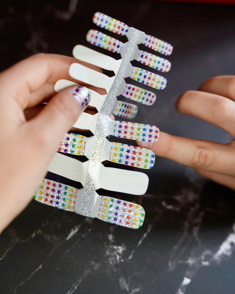 Kinderhand hält bunte Glitzer-Nagelfolien mit Regenbogen-Sternchen von SHEESH Nails – fröhliches Naildesign für Kinder mit funkelndem Regenbogenmuster.