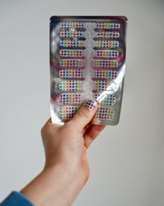 Kinderhand hält bunte Glitzer-Nagelfolien mit Regenbogen-Sternchen von SHEESH Nails – fröhliches Naildesign für Kinder mit funkelndem Regenbogenmuster.