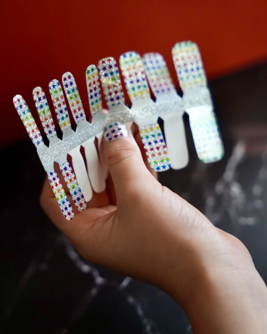 Kinderhand hält bunte Glitzer-Nagelfolien mit Regenbogen-Sternchen von SHEESH Nails – fröhliches Naildesign für Kinder mit funkelndem Regenbogenmuster.