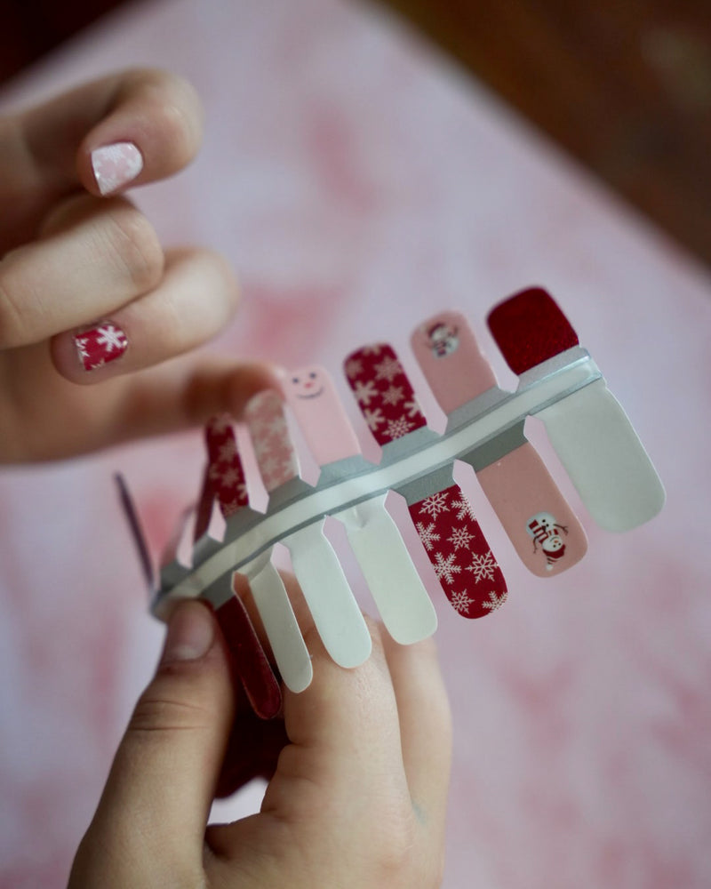 Kinderhand appliziert rote und rosafarbene Kinder-Nagelfolien mit Schneemann- und Schneeflocken-Motiv von SHEESH Nails – weihnachtliches Winter Nail Design für Kinderhände.
