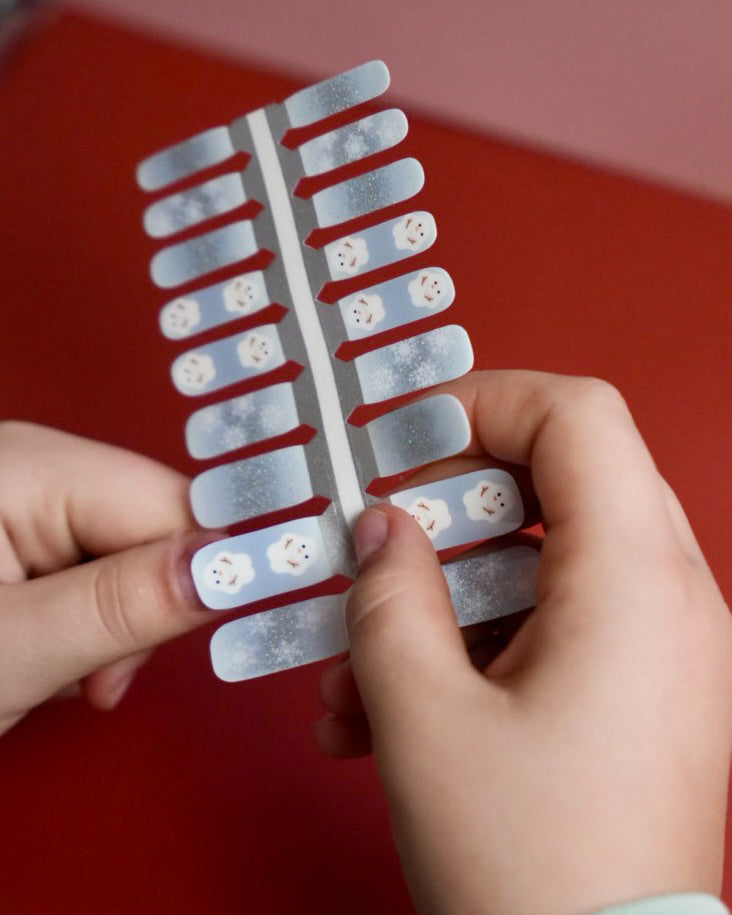 Kinderhand appliziert blaue Schneemann Nagelfolien von SHEESH auf rotem Hintergrund  winterliches Nageldesign für Kinder mit Schneemann-Motiv. 