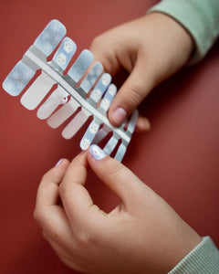 Kinderhand appliziert blaue Schneemann Nagelfolien von SHEESH auf rotem Hintergrund  winterliches Nageldesign für Kinder mit Schneemann-Motiv. 