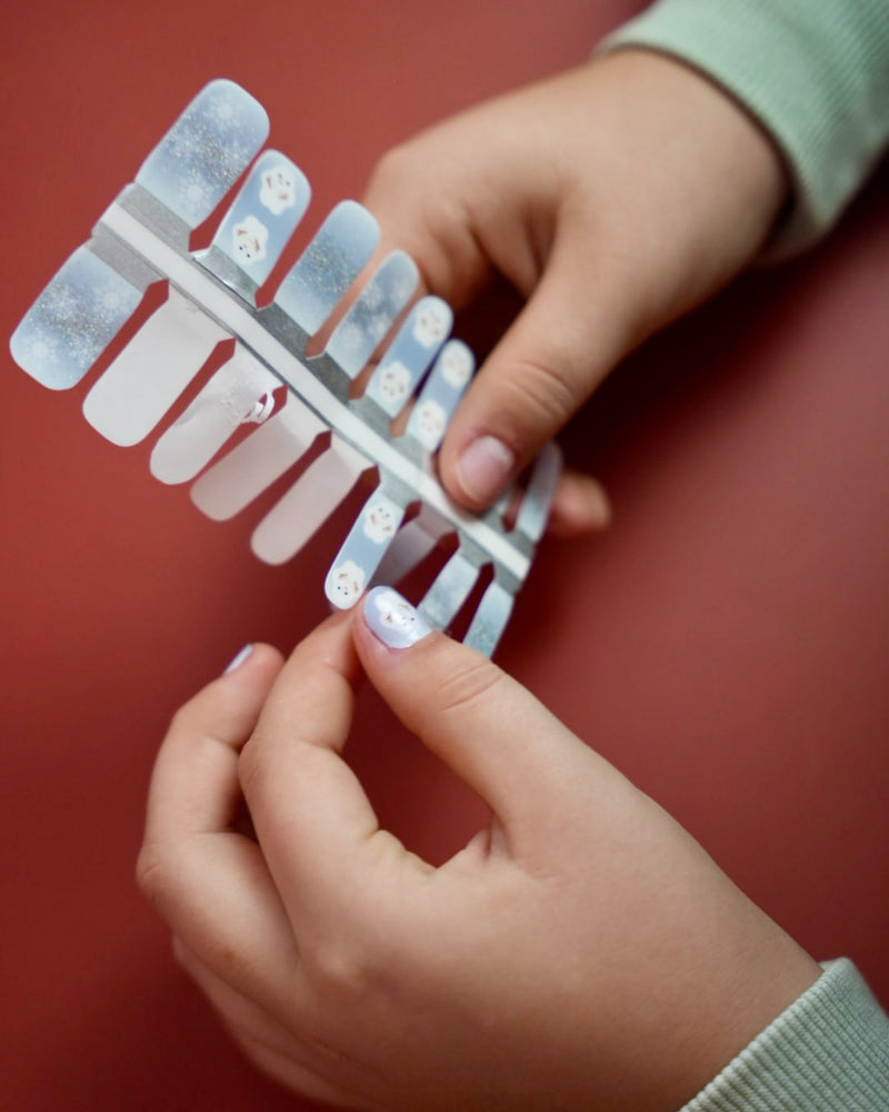 Kinderhand appliziert blaue Schneemann Nagelfolien von SHEESH auf rotem Hintergrund  winterliches Nageldesign für Kinder mit Schneemann-Motiv. 