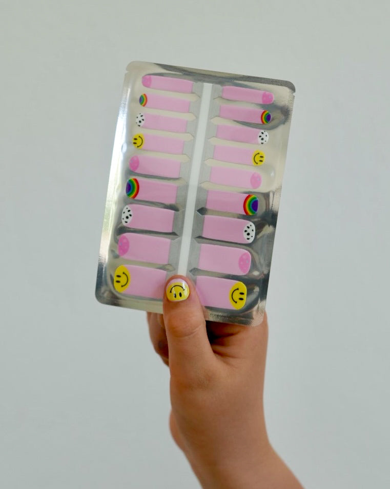 Nahaufnahme einer Kinderhand mit den Sheesh Nails Kinder-Nagelfolien „Smiley“. Die selbstklebenden Sticker zeigen ein fröhliches Design mit pinkem Hintergrund und gelben Smiley-Motiven. Das deckende Finish sorgt für einen bunten, kindgerechten Look. Hautfreundlich, ungiftig und einfach anzubringen – ideal für kreative Kinder-Maniküren voller Spaß und Farbe.