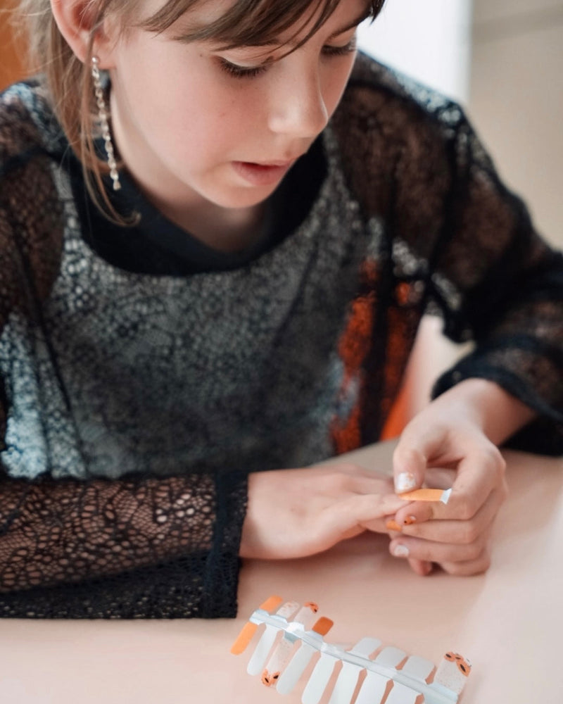 Nahaufnahme einer Kinderhand mit Sheesh Nails Kinder-Nagelfolien „Sonnenblume“. Die selbstklebenden Sticker zeigen ein fröhliches Design mit orangefarbenen und weißen Streifen sowie kleinen Sonnenblumenmotiven. Das verspielte Muster sorgt für einen sommerlichen, kindgerechten Look. Die Folien sind hautfreundlich, ungiftig und leicht anzubringen – ideal für kreative Kinder-Maniküren mit Blumenmotiven.