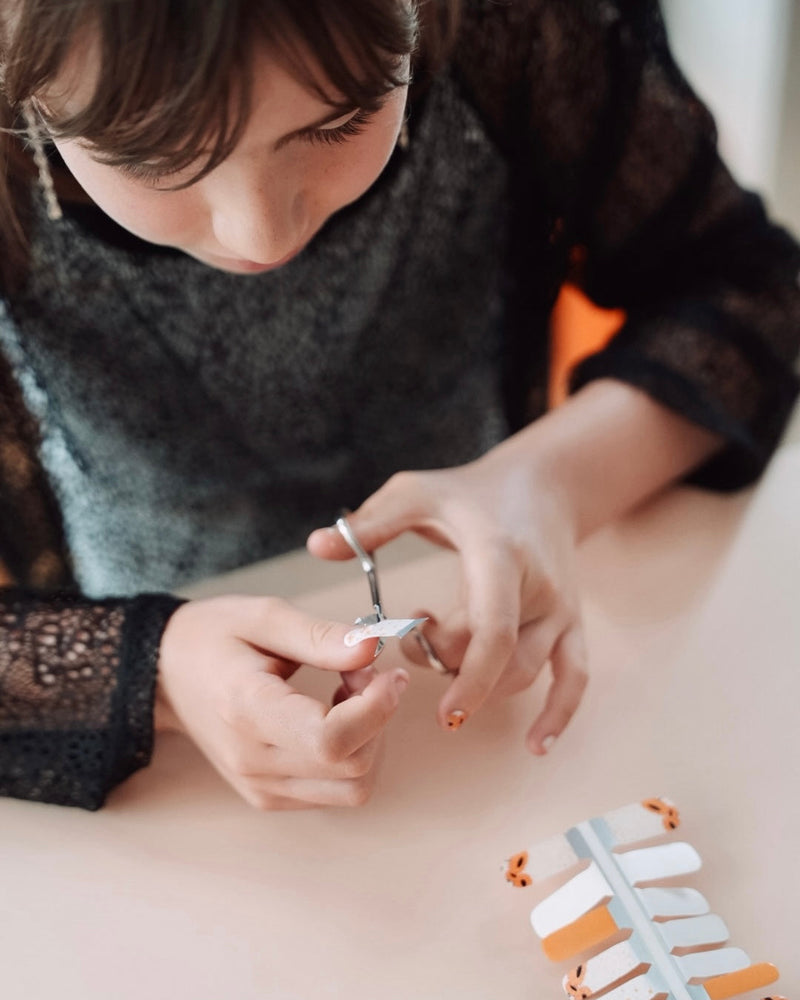 Nahaufnahme einer Kinderhand mit Sheesh Nails Kinder-Nagelfolien „Sonnenblume“. Die selbstklebenden Sticker zeigen ein fröhliches Design mit orangefarbenen und weißen Streifen sowie kleinen Sonnenblumenmotiven. Das verspielte Muster sorgt für einen sommerlichen, kindgerechten Look. Die Folien sind hautfreundlich, ungiftig und leicht anzubringen – ideal für kreative Kinder-Maniküren mit Blumenmotiven.