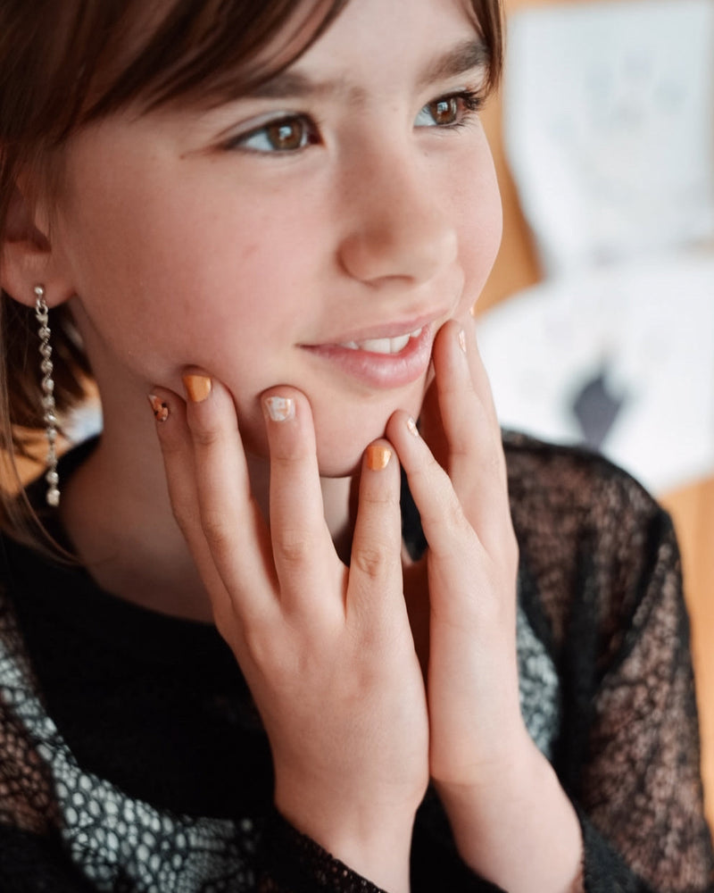 Nahaufnahme einer Kinderhand mit Sheesh Nails Kinder-Nagelfolien „Sonnenblume“. Die selbstklebenden Sticker zeigen ein fröhliches Design mit orangefarbenen und weißen Streifen sowie kleinen Sonnenblumenmotiven. Das verspielte Muster sorgt für einen sommerlichen, kindgerechten Look. Die Folien sind hautfreundlich, ungiftig und leicht anzubringen – ideal für kreative Kinder-Maniküren mit Blumenmotiven.