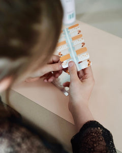 Nahaufnahme einer Kinderhand mit Sheesh Nails Kinder-Nagelfolien „Sonnenblume“. Die selbstklebenden Sticker zeigen ein fröhliches Design mit orangefarbenen und weißen Streifen sowie kleinen Sonnenblumenmotiven. Das verspielte Muster sorgt für einen sommerlichen, kindgerechten Look. Die Folien sind hautfreundlich, ungiftig und leicht anzubringen – ideal für kreative Kinder-Maniküren mit Blumenmotiven.
