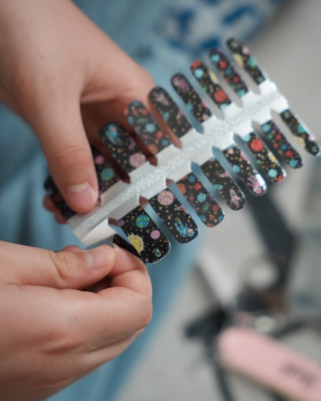 Nahaufnahme einer Kinderhand mit den Sheesh Nails Kinder-Nagelfolien „Space“. Die selbstklebenden Sticker zeigen ein galaktisches Design mit bunten Planeten und Sternen auf schwarzem Hintergrund. Das deckende Finish sorgt für einen kräftigen, farbenfrohen Look. Hautfreundlich, ungiftig und einfach anzubringen – ideal für kreative Kinder-Maniküren mit Weltraum-Motiven.