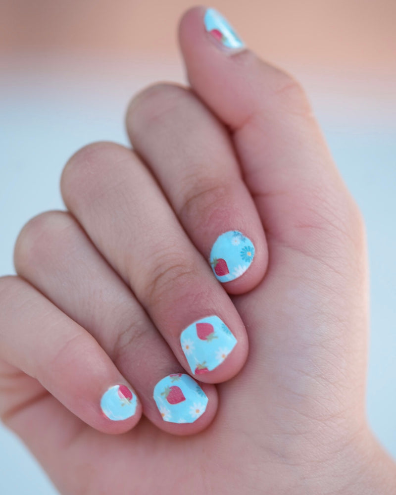 Nahaufnahme einer Kinderhand mit den Sheesh Nails Kinder-Nagelfolien „Süßes Früchtchen“. Die selbstklebenden Sticker zeigen ein sommerlich-verspieltes Design mit roten Erdbeeren auf hellblauem Grund. Das fröhliche Motiv sorgt für gute Laune und bringt Farbe in jede Kinder-Maniküre. Die Folien sind deckend, leicht anzubringen und rückstandsfrei wieder zu entfernen – ganz ohne UV-Lampe. Hautfreundlich, ungiftig und perfekt für kreative kleine Nail-Artists, die Spaß an bunten Designs haben.