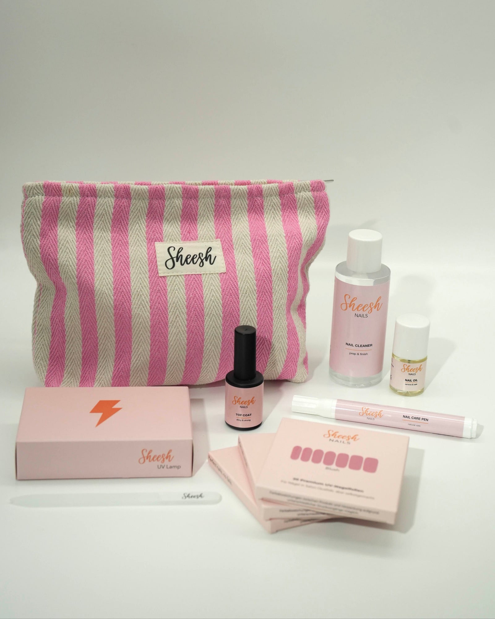 Produktfoto des Sheesh Nails Nagelfolien Starter-Sets mit Beauty Bag. Enthalten sind UV-Lampe, Top Coat, Nail Cleanser, Remover, Feile und Nagelfolien – alles, was für eine vollständige Maniküre benötigt wird. Die rosa-weiß gestreifte Sheesh Beauty Bag sorgt für Ordnung und Stil. Perfekt für Einsteigerinnen, die zu Hause professionelle Ergebnisse erzielen möchten.