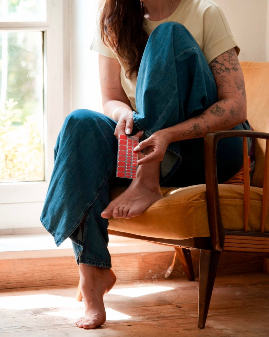 Frau bringt die Sheesh Nails Pedi UV-Nagelfolie „Lifeguard“ auf ihren Zehennägeln an. In entspannter Atmosphäre sitzt sie am Fenster und platziert die leuchtend rote Folie präzise auf dem Nagel. Das natürliche Licht betont die sommerliche Farbintensität und vermittelt einen gepflegten, modernen Look für zu Hause.