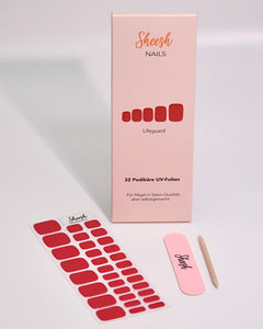Produktaufnahme der Sheesh Nails Pedi UV-Nagelfolie „Lifeguard“. Die Verpackung präsentiert das kräftige Rot der Folien, kombiniert mit Feile und Holzstäbchen. Das minimalistische Setup und das helle Licht betonen die hochwertige Qualität und den modernen, gepflegten Salon-Look – ideal für elegante Sommerpediküren mit leuchtendem Finish.