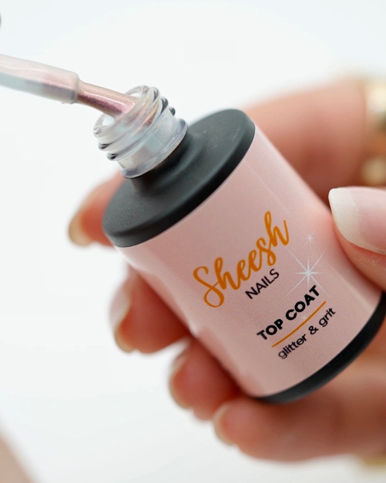 Produktfotografie des Sheesh Nails Top Coat „Glitter & Grit“. Die geöffnete Flasche zeigt den Pinsel mit schimmerndem Versiegelungslack in zartem Rosaton. Der transparente Top Coat sorgt für langanhaltenden Glanz, schützt die UV-Nagelfolien und verleiht ein professionelles, glänzendes Finish.