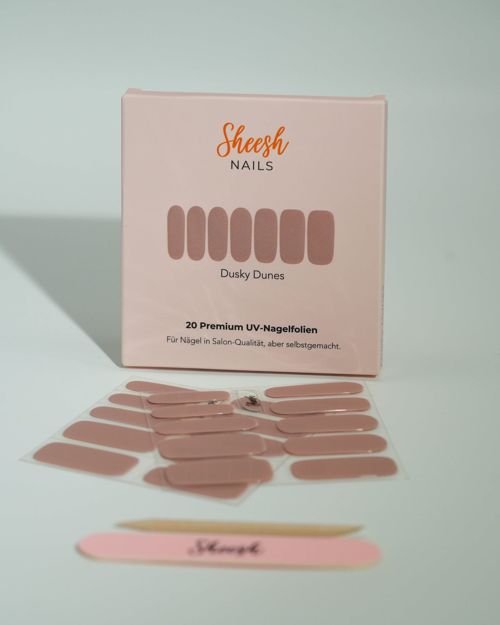 Produktfoto der Sheesh Nails UV-Nagelfolien „Dusky Dunes“. Die Folien in elegantem Nude-Ton bieten ein dezentes, glänzendes Finish für gepflegte Nägel in Salonqualität. Im Vordergrund liegen geöffnete Folien und eine Sheesh-Nagelfeile – stilvoll präsentiert und ideal für natürliche, zeitlose Maniküren