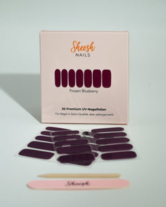 Produktfoto der Sheesh Nails UV-Nagelfolien „Frozen Blueberry“. Die selbstklebenden Folien in tiefem Lila mit kühlem Glanz verleihen den Nägeln einen edlen, modernen Look in Salonqualität. Im Vordergrund liegen die geöffneten Folien und eine Sheesh-Nagelfeile – ideal für elegante, ausdrucksstarke Maniküren mit winterlichem Charakter.