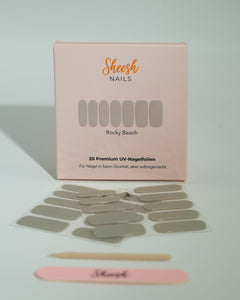 Produktfoto der Sheesh Nails UV-Nagelfolien „Rocky Beach“. Die selbstklebenden Folien in elegantem Beige verleihen den Nägeln einen natürlichen, glänzenden Look in Salonqualität. Im Vordergrund liegen die geöffneten Folien und eine Sheesh-Nagelfeile – ideal für dezente, gepflegte Maniküren mit modernem Touch.