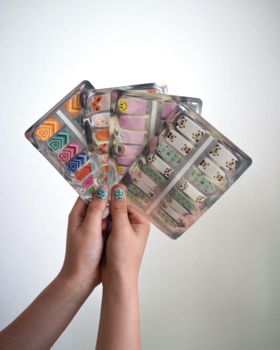 SHEESH Nails – Space | Kinder-Nagelfolien, Bild 06: Nahaufnahme von Kinderhänden, die mehrere Kinder-Nagelfolien-Verpackungen mit verschiedenen Designs halten.