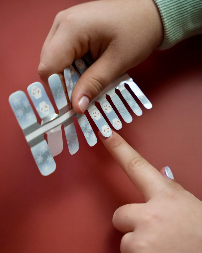 Kinderhand appliziert blaue Schneemann Nagelfolien von SHEESH auf rotem Hintergrund  winterliches Nageldesign für Kinder mit Schneemann-Motiv. 